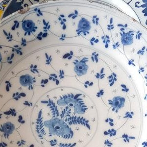 May include: A collection of antique white ceramic plates with blue floral patterns. The plates feature intricate designs of flowers and vines, with a central floral motif. The plates are arranged on a colorful patterned tablecloth.