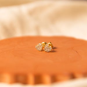 May include: A pair of gold heart-shaped stud earrings, each set with small, sparkling stones. The earrings are presented on a reddish-brown, irregularly shaped surface, with a soft, off-white fabric in the background. The earrings are small and delicate.