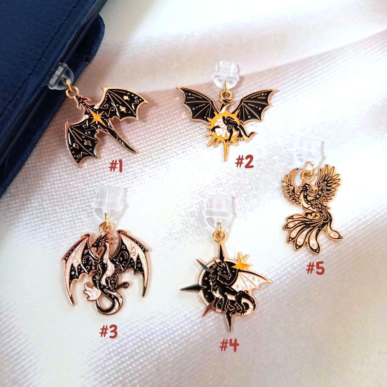 May include: Five gold-toned metal charms featuring black enamel dragon and phoenix designs, each labeled with a number from #1 to #5. The charms include celestial motifs like stars and moons, and each is attached to a clear plastic earring back.