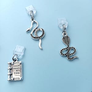 Peut inclure: Trois boucles d'oreilles argent&eacute;es sur fond bleu clair. Deux pr&eacute;sentent des motifs de serpents, l'un enroul&eacute; et l'autre avec une t&ecirc;te de cobra. La troisi&egrave;me boucle d'oreille est un petit livre avec le texte "Once Upon A Time".
