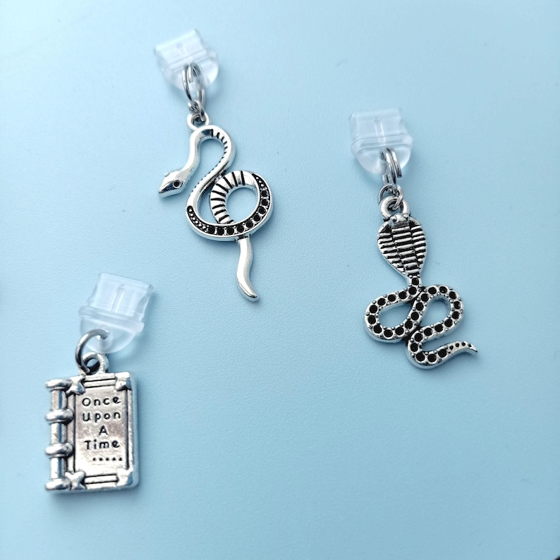 Peut inclure: Trois boucles d'oreilles argent&eacute;es sur fond bleu clair. Deux pr&eacute;sentent des motifs de serpents, l'un enroul&eacute; et l'autre avec une t&ecirc;te de cobra. La troisi&egrave;me boucle d'oreille est un petit livre avec le texte "Once Upon A Time".
