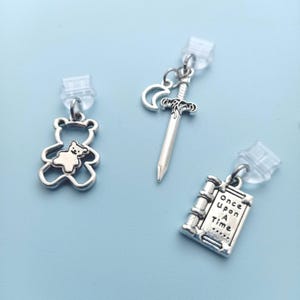 Peut inclure: Boucles d'oreilles &agrave; breloques argent&eacute;es avec un ours en peluche, une &eacute;p&eacute;e et un croissant de lune, et un livre avec le texte "Once Upon A Time". Les breloques sont fix&eacute;es &agrave; des clous d'oreilles transparents.