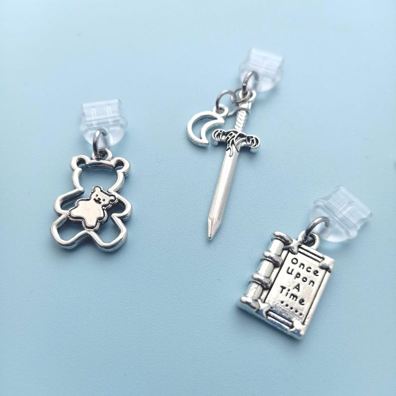 Peut inclure: Boucles d'oreilles &agrave; breloques argent&eacute;es avec un ours en peluche, une &eacute;p&eacute;e et un croissant de lune, et un livre avec le texte "Once Upon A Time". Les breloques sont fix&eacute;es &agrave; des clous d'oreilles transparents.