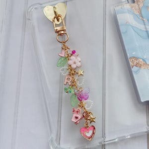 Op de afbeelding: Een goudkleurige telefoonbedel met een hartvormige sluiting en een bungelende ketting. De ketting heeft roze en gouden bedels, waaronder een hart, sterren, vlinders, bloemen en de letter "R". De bedel is bevestigd aan een doorzichtige telefoonhoes.