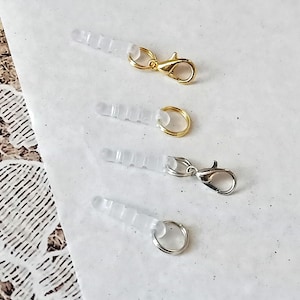 Può includere: Quattro tappi antipolvere trasparenti per auricolari con dettagli metallici. Due hanno chiusure e anelli color oro, mentre gli altri due hanno chiusure e anelli color argento. I tappi sono disposti su una superficie bianca.