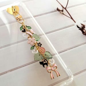 Handmade botanical charm chain clipped to a clear Kindle case, featuring gold heart clasp, pastel bow, flower and leaf charms.