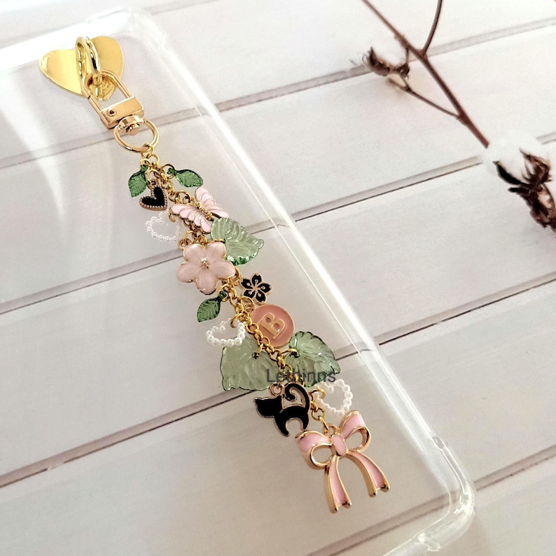 Handmade botanical charm chain clipped to a clear Kindle case, featuring gold heart clasp, pastel bow, flower and leaf charms.