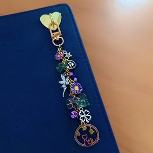 May include: A gold-toned metal keychain featuring a heart-shaped clasp and a variety of charms including a silver fairy, purple and yellow flowers, green leaves, a four-leaf clover, a star, and a circular pendant with a cat silhouette.