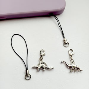 Puede incluir: Encantos de teléfono de dinosaurio plateados con cordones negros. Un dije es un estegosaurio, el otro es un brontosaurio. Los dijes están unidos a un cordón negro con un anillo plateado. Un teléfono morado está en el fondo.