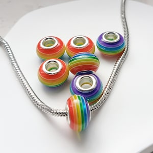 Puede incluir: Un conjunto de cuentas a rayas de arcoíris con detalles metálicos plateados. Las cuentas están ensartadas en una cadena plateada, mostrando una vibrante gama de colores. Las cuentas son redondas y tienen un acabado brillante, perfectas para bisutería.