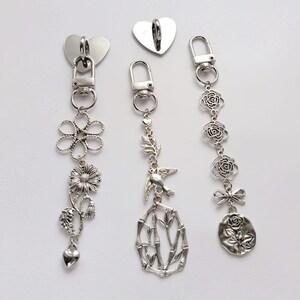 Könnte beinhalten: Silberfarbene Metallanhänger mit verschiedenen Designs, darunter ein Herz, eine Blume, ein Vogel mit Bambus und Rosen. Jeder Anhänger hat einen Verschluss zur Befestigung. Die Anhänger werden vor weißem Hintergrund gezeigt.