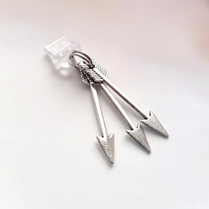 May include: Silver-toned arrow earrings. Each earring features three arrows with detailed fletching and pointed tips. The earrings are attached to a clear earring back. The arrows are a light silver color.