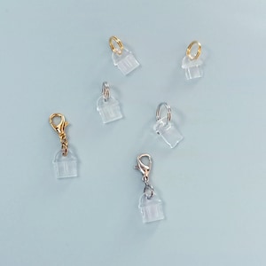 Könnte beinhalten: Eine Sammlung klarer Kunststoffanhänger mit Metallverschlüssen und Ringen. Die Verschlüsse sind gold-, silber- und roségoldfarben. Die Anhänger sind wie kleine Häuser oder Gebäude geformt, auf hellblauem Hintergrund.