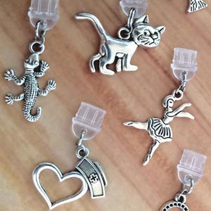 Könnte beinhalten: Ohrringe mit silberfarbenen Anhängern in verschiedenen Designs. Die Kollektion umfasst eine Eidechse, eine Katze, eine Ballerina, ein Herz mit einem medizinischen Symbol und ein kreisförmiges Design. Jeder Anhänger ist an einem transparenten Ohrstecker befestigt.