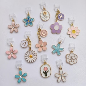 Pode incluir: Uma coleção de pingentes coloridos em forma de flor com detalhes em metal dourado. Cada pingente apresenta um design diferente, incluindo margaridas, rosas e letras iniciais. Presos a clipes de plástico transparente.