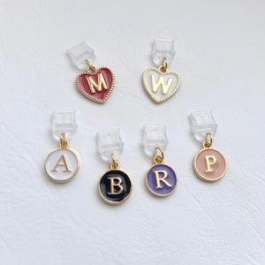 Könnte beinhalten: Sechs kleine Anhänger mit goldfarbenen Metallrändern und durchsichtigen Kunststoffbefestigungen. Zwei sind herzförmig mit roter und weißer Emaille und den Buchstaben M und W. Vier sind kreisförmig mit weißer, schwarzer, violetter und rosa Emaille mit A, B, R, P.