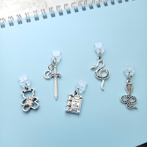 Puede incluir: Cinco colgantes plateados con clips de plástico transparente. Los colgantes incluyen un oso de peluche, una espada, un libro con el texto "Once Upon a Time" y dos diseños de serpiente. Sobre una superficie azul claro.