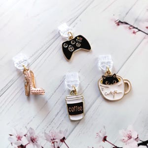Könnte beinhalten: Eine Sammlung von Clip-on-Anhängern, darunter ein High-Heel-Schuh, ein Videospiel-Controller, eine Kaffeetasse mit dem Wort "coffee" und eine Katze in einer Tasse. Die Anhänger sind goldfarben mit schwarzen, weißen und rosa Akzenten.