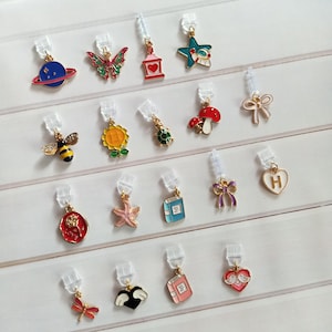 Puede incluir: Una colección de pendientes de clip con varios dijes. Los diseños incluyen un planeta, mariposa, linterna, estrella, abeja, girasol, tortuga, hongo, lazo, corazón, rosa, estrella de mar, libro, libélula, corazón alado y un corazón con pájaros.