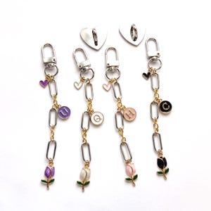May include: Four keychains with silver-tone clasps, linked chains, and decorative charms. Each keychain features a different colored tulip charm, a heart charm, and a charm with a letter. Two silver heart-shaped charms are also visible.