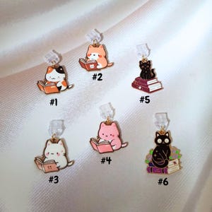 May include: Six cat-themed earrings are displayed. Each earring showcases a different coloured cat reading a book. The cats are in various colours, including orange, black, white, and pink. The earrings are attached to clear earring posts.
