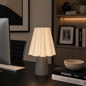May include: A contemporary desk lamp featuring a pleated white lampshade and a grey concrete base. The lamp is lit, showcasing the shade's design. It rests on a black desk alongside books and a decorative bowl. The background includes a bookshelf and artwork.