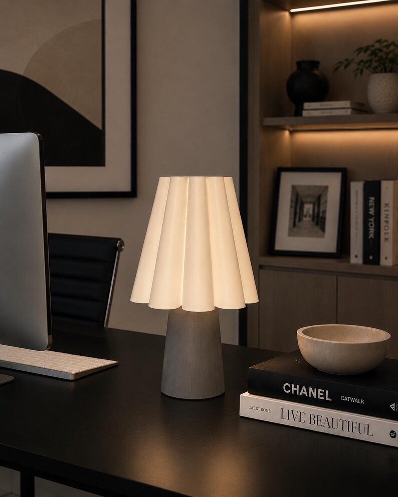 May include: A contemporary desk lamp featuring a pleated white lampshade and a grey concrete base. The lamp is lit, showcasing the shade's design. It rests on a black desk alongside books and a decorative bowl. The background includes a bookshelf and artwork.