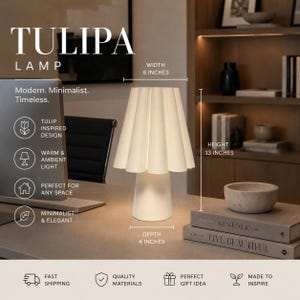 May include: A white Tulip lamp with a pleated shade, 33 cm tall, 20 cm wide, and 10 cm deep. The lamp is on a desk with books and a bowl. Text on the lamp reads "TULIPA LAMP".
