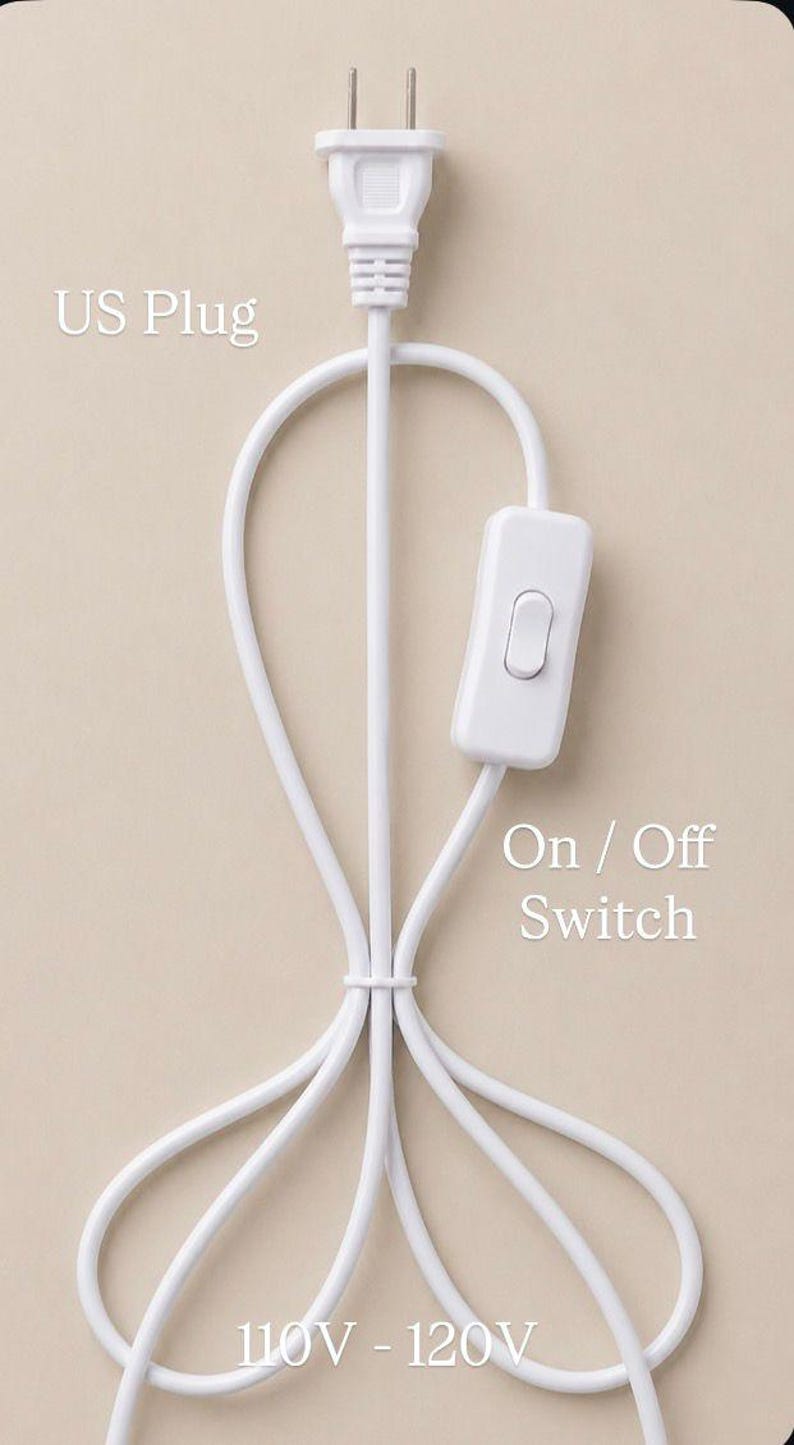 May include: A white electrical cord featuring a US plug, an on/off switch, and the text "110V - 120V". The cord is looped and tied together. The background is a plain beige colour.