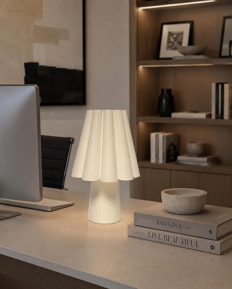 May include: A white table lamp with a pleated shade is on a desk. The lamp has a rounded base and a flared shade. The desk also holds books, a marble bowl, and a computer monitor. The background includes a bookshelf and artwork.