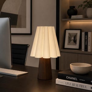 May include: A contemporary table lamp featuring a white, pleated lampshade and a dark wooden base. The lamp sits on a dark desk alongside books and a small bowl. The background includes a computer monitor, artwork, and a bookshelf.
