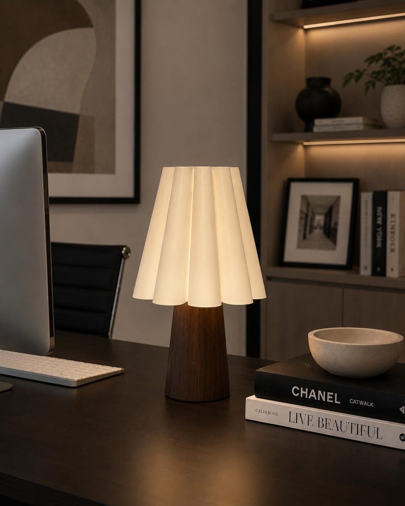May include: A contemporary table lamp featuring a white, pleated lampshade and a dark wooden base. The lamp sits on a dark desk alongside books and a small bowl. The background includes a computer monitor, artwork, and a bookshelf.