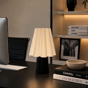 May include: A stylish desk lamp featuring a pleated white lampshade and a black base. The lamp is positioned on a black desk, alongside a computer monitor and books. The books include titles such as "CHANEL" and "LIVE BEAUTIFUL".