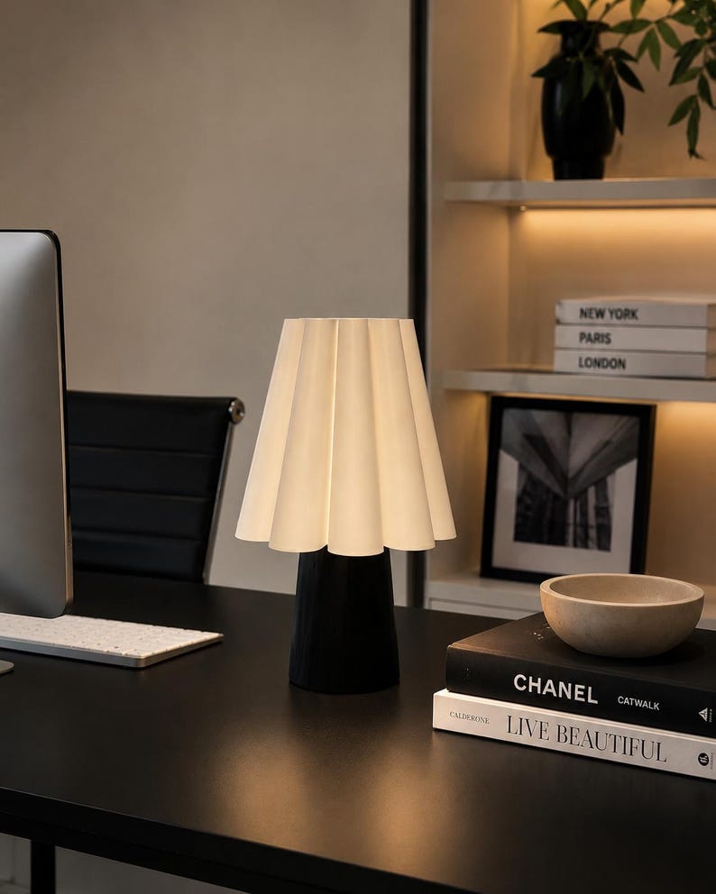 May include: A stylish desk lamp featuring a pleated white lampshade and a black base. The lamp is positioned on a black desk, alongside a computer monitor and books. The books include titles such as "CHANEL" and "LIVE BEAUTIFUL".