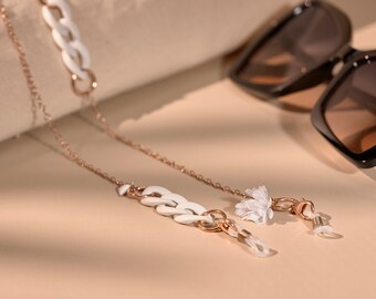 May include: A rose gold chain with white links and a delicate white flower charm, designed to hold eyeglasses. Black sunglasses with brown lenses are in the background. The chain is on a beige surface.