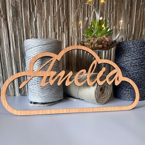 May include: Wooden cloud-shaped sign with the name "Amelia" cut out in a cursive font. The sign is sitting on top of two spools of twine, one gray and one beige.