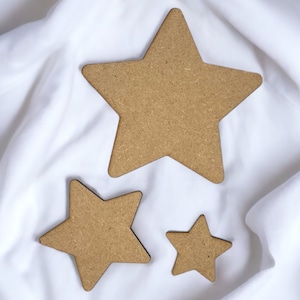 Op de afbeelding: Drie houten sterren van verschillende groottes op een witte stoffen achtergrond. De grootste ster bevindt zich in het midden, met een kleinere ster links en de kleinste ster rechts.