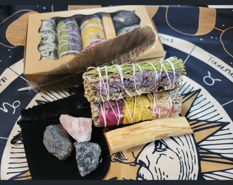 Può includere: Una collezione di articoli spirituali tra cui mazzi di salvia, cristalli e palo santo. I mazzi di salvia sono avvolti con spago e adornati con fiori secchi. Un sacchetto di velluto nero contiene diversi cristalli grezzi. Una grande piuma è appoggiata vicino ai mazzi.