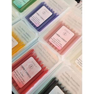 Könnte beinhalten: Eine Auswahl an Glockenkerzen in verschiedenen Farben, darunter Rot, Rosa, Lila und Grün, verpackt in transparenten Kunststoffbehältern. Jede Packung hat ein Etikett mit dem Markennamen "OTHALA" und dem Text "CHIME CANDLES". Die durchschnittliche Brenndauer beträgt 2-2,5 Stunden.