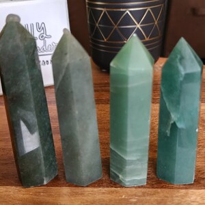 May include: Four green crystal obelisks of varying shades and textures stand on a wooden surface. The obelisks are faceted and have pointed tops. The crystals range in color from a deep forest green to a lighter, translucent green.