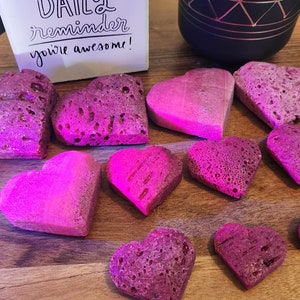 May include: A collection of heart-shaped objects in varying shades of pink and purple, arranged on a wooden surface. Some hearts have a porous texture, while others are smooth. A small card with the text "DAILY reminder you're awesome!" is also present.
