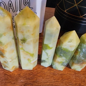 May include: Five yellow and green crystal points, also known as wands, are arranged in a row on a wooden surface. The crystals have a marbled pattern of yellow, green, and white.