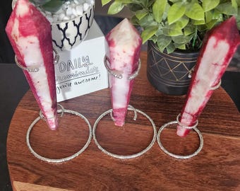 May include: Three pink and white crystal wands with silver metal stands on a wooden surface. The wands are arranged in a row, with the largest wand on the left and the smallest wand on the right. The wands are all pointed at the top and have a smooth, polished finish.