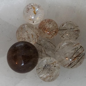 May include: A collection of clear quartz spheres with internal inclusions of black and gold rutile needles. One sphere is a darker, smoky quartz. The spheres vary in size and are arranged on a white surface. These gemstones are ideal for jewelry making or crystal healing.