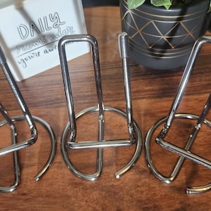 May include: Four silver metal picture stands with a circular base. The stands are arranged in a row on a wooden surface.