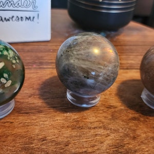 May include: Three polished gemstone spheres on clear stands. The spheres are green with white flecks, gray with iridescent shimmer, and brown with white streaks.