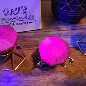 May include: Three pink geometric shapes on gold metal stands. The stands are triangular and have a geometric design. The shapes are on a wooden surface.