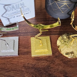May include: Three decorative metal stands for displaying jewelry or other small items. The stands are made of gold-colored metal and have different base designs. One stand has a square base, another has a round base with a decorative pattern, and the third has a square base with a textured surface.