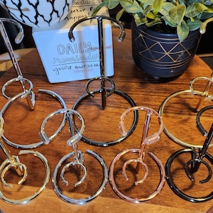 May include: Six metal stands for displaying mugs or cups. The stands are made of different metals, including gold, silver, and rose gold. The stands are arranged in a circle on a wooden surface.