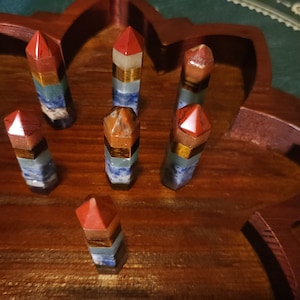 May include: Seven multi-colored crystal points arranged in a wooden tray. The points are made of different types of crystals, including amethyst, clear quartz, tiger's eye, and red jasper. The tray is carved in the shape of a flower.